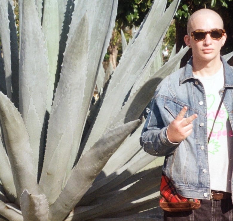 A person wearing a denim jacket makes a peace sign while standing next to a large cactus.