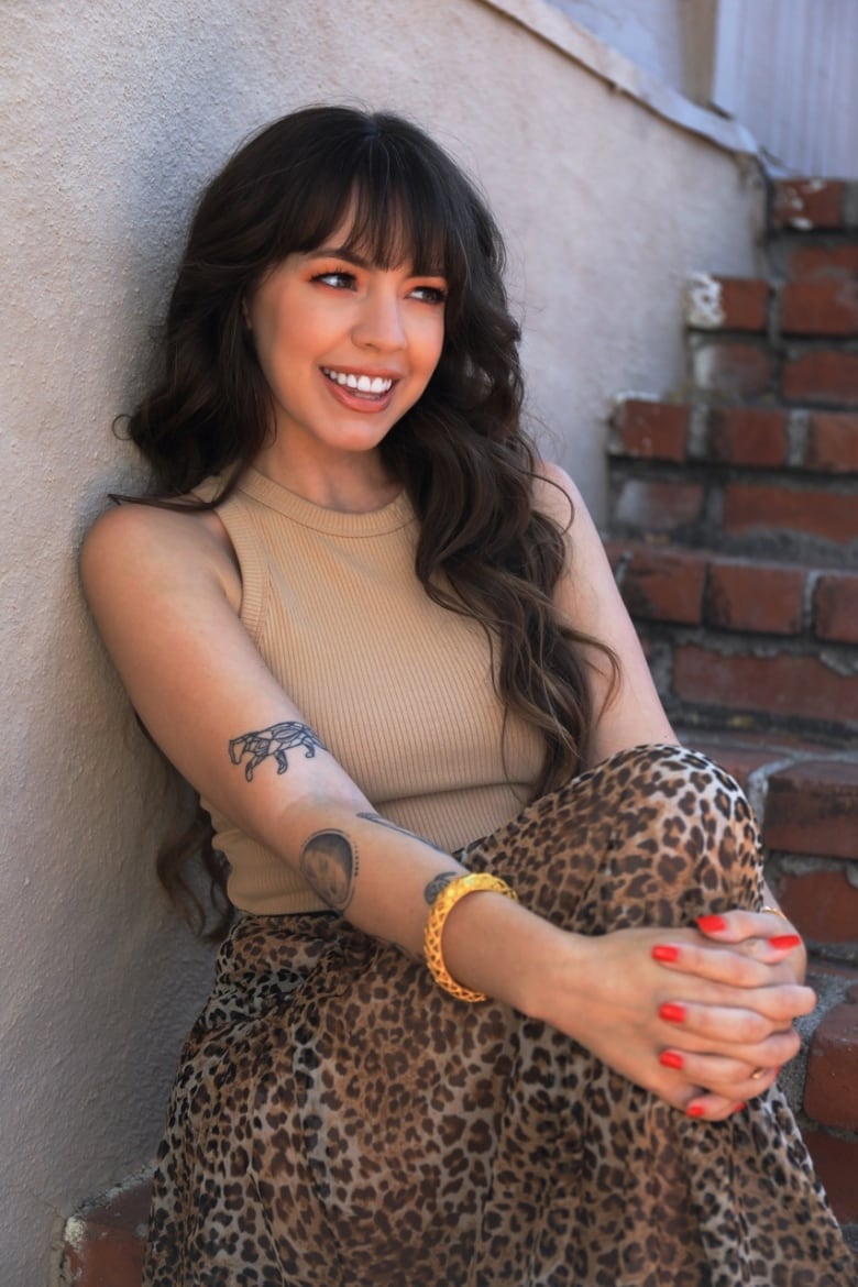 A smiling woman with long brown hair sits on a brick outdoor step with her hands wrapped around one knee.